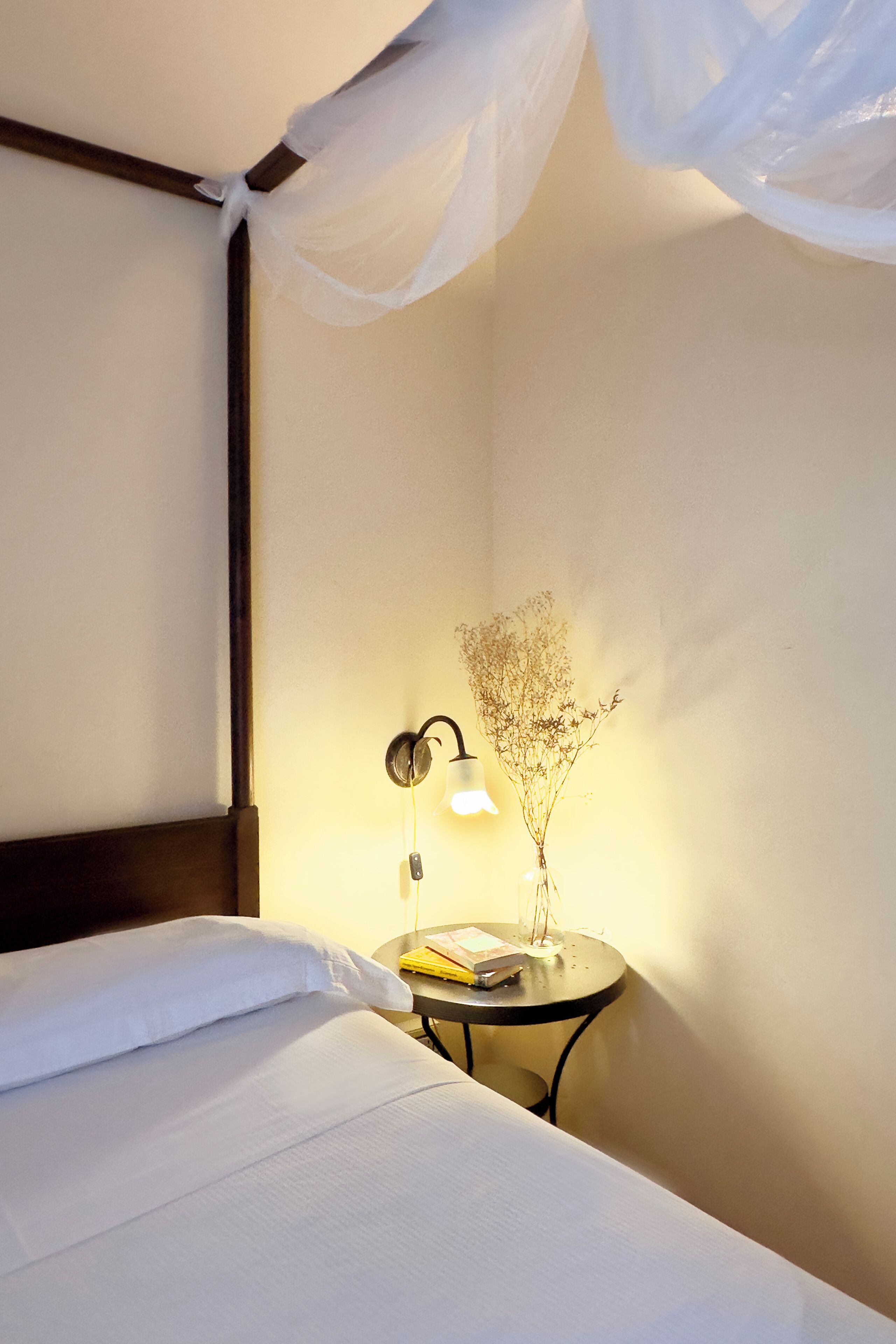 A neatly made bed with white linens, a bedside lamp, and a small table with a vase of dried flowers.