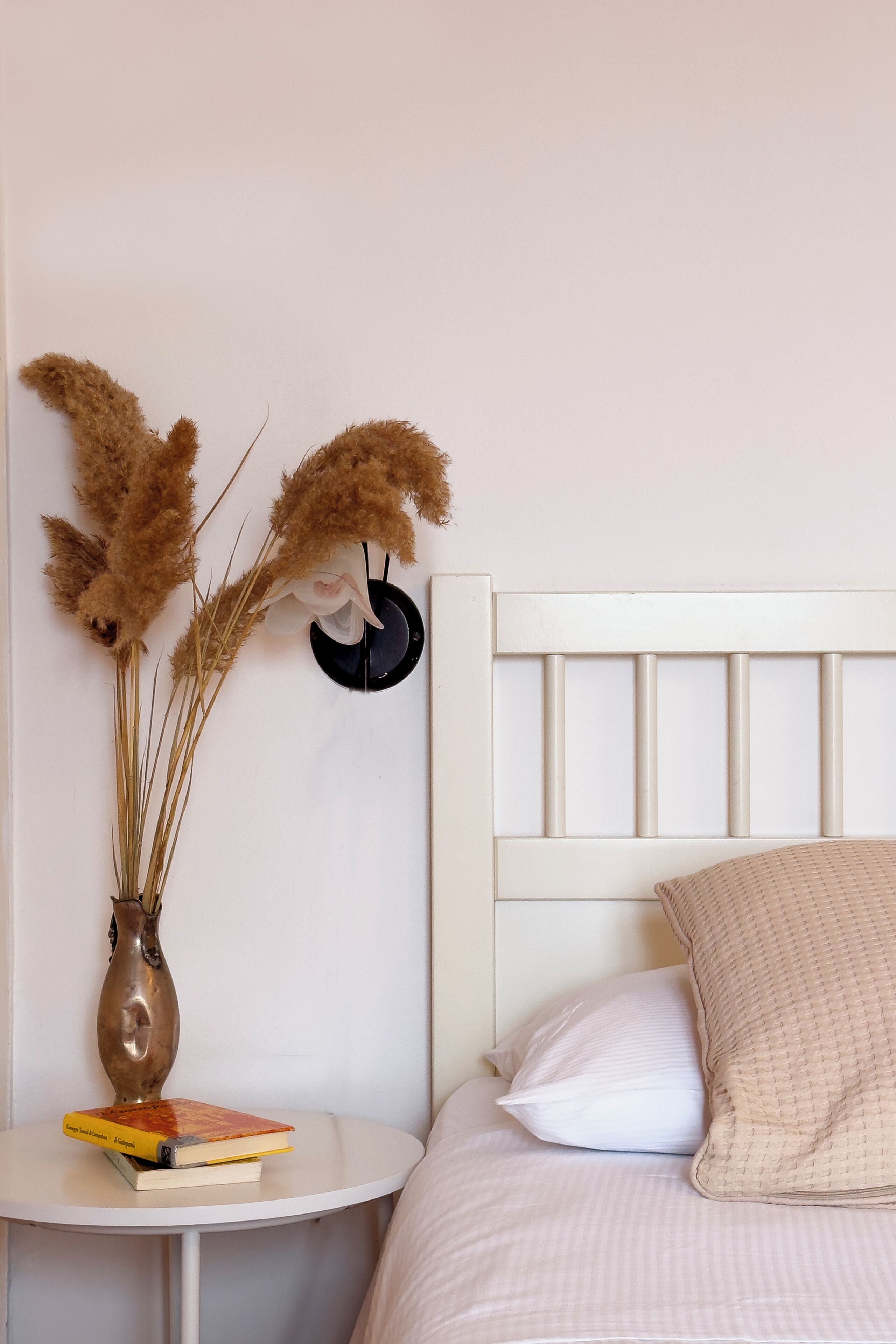 A neatly arranged bedroom with a white bed, a bedside table with a book and a vase of dried pampas grass, and a wall-mounted black object.