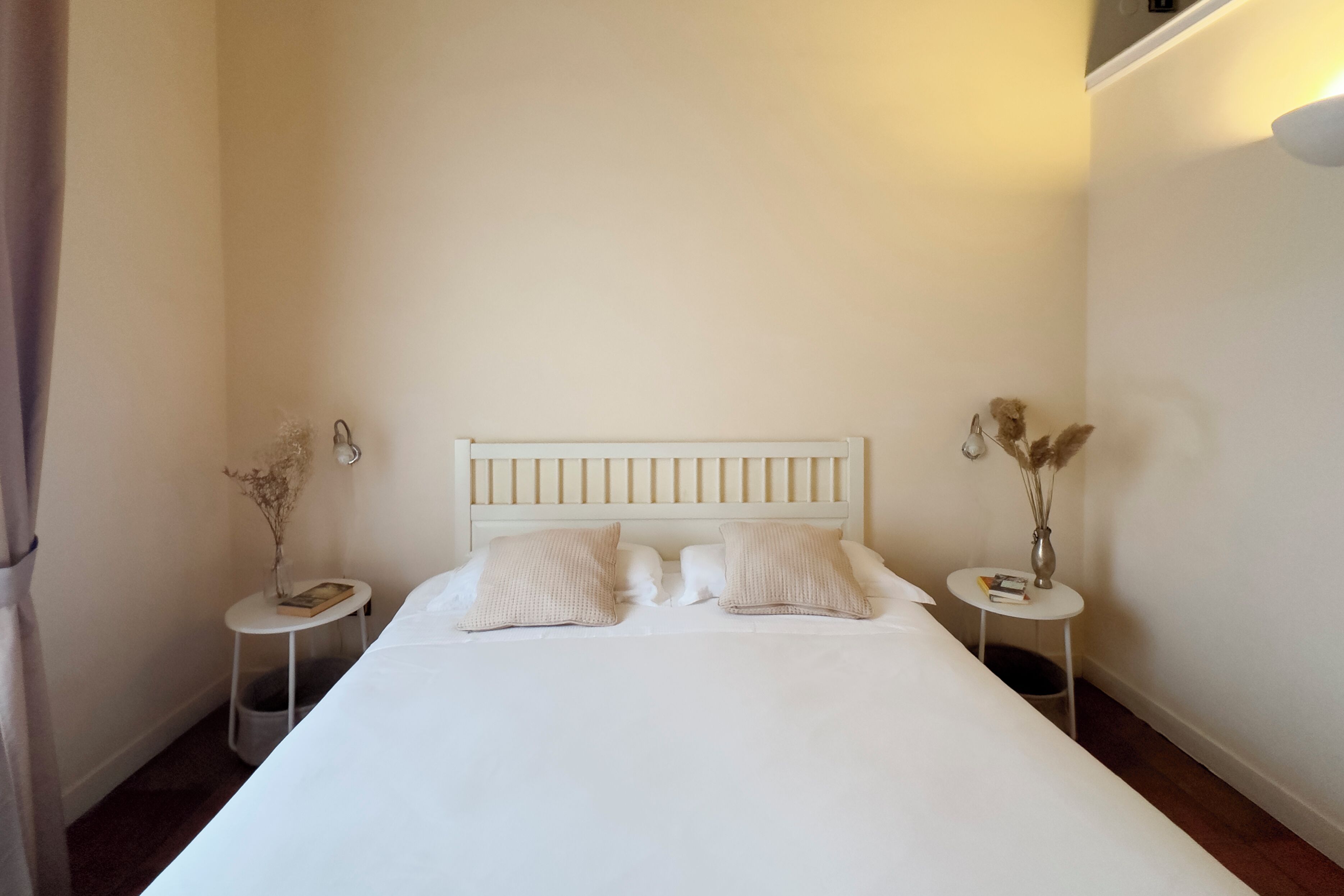 Una cama bien hecha con ropa blanca y dos almohadas, flanqueada por dos mesitas de noche pequeñas, blancas, con libros y un jarrón de flores secas.
