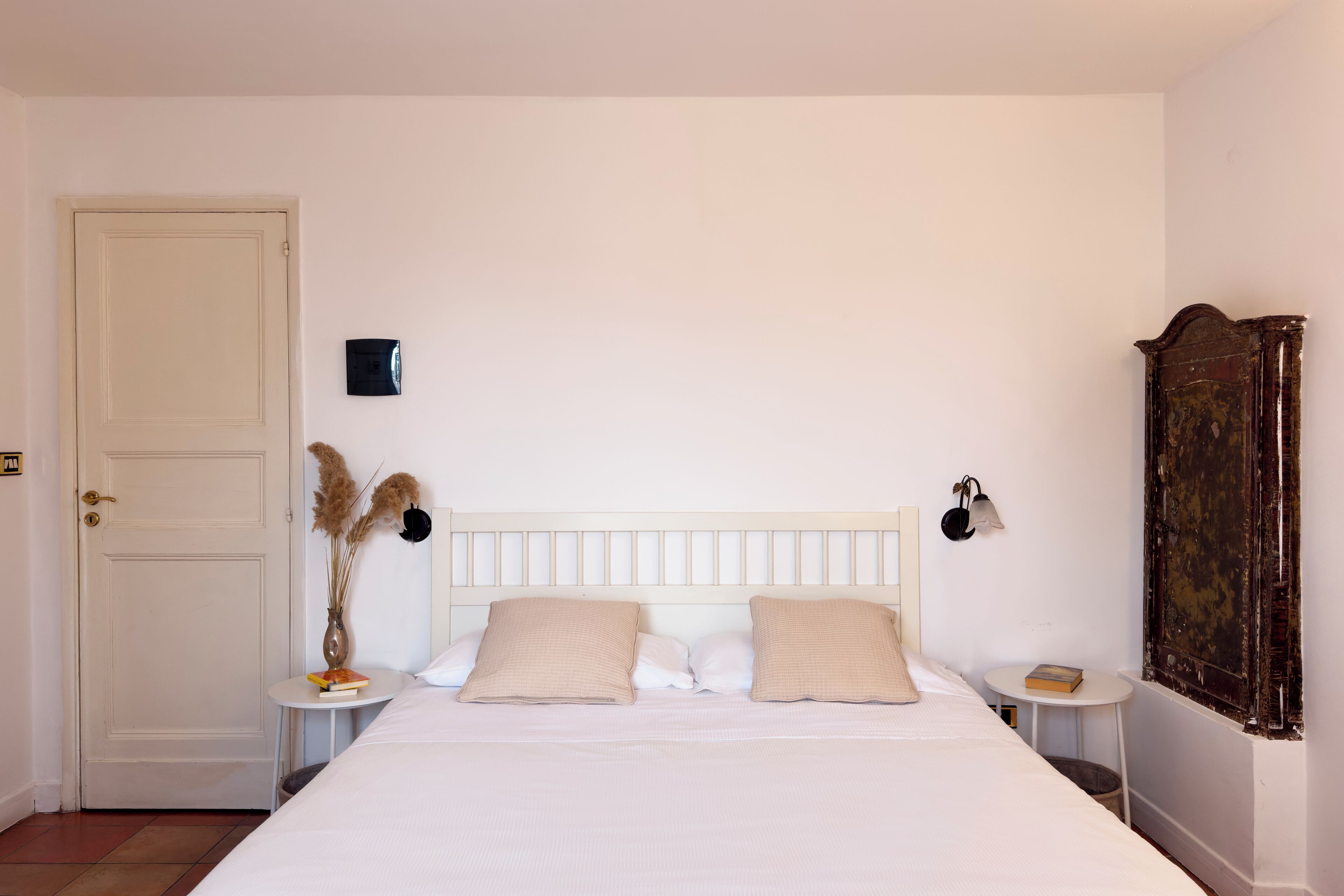 A bedroom with a bed, two bedside tables, a nightstand, a door, a wall-mounted light, a small table with a book, and a window with a curtain.