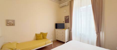 A bedroom with a single bed, a bedside table with a TV, a dresser, and a window with curtains.