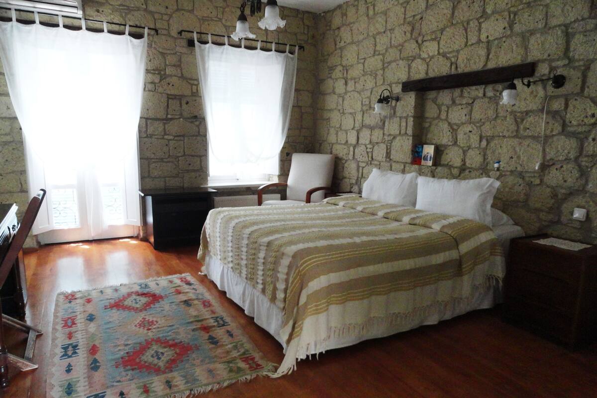 A bedroom with a stone wall, a bed with a striped bedspread, a chair, a desk, and a rug.