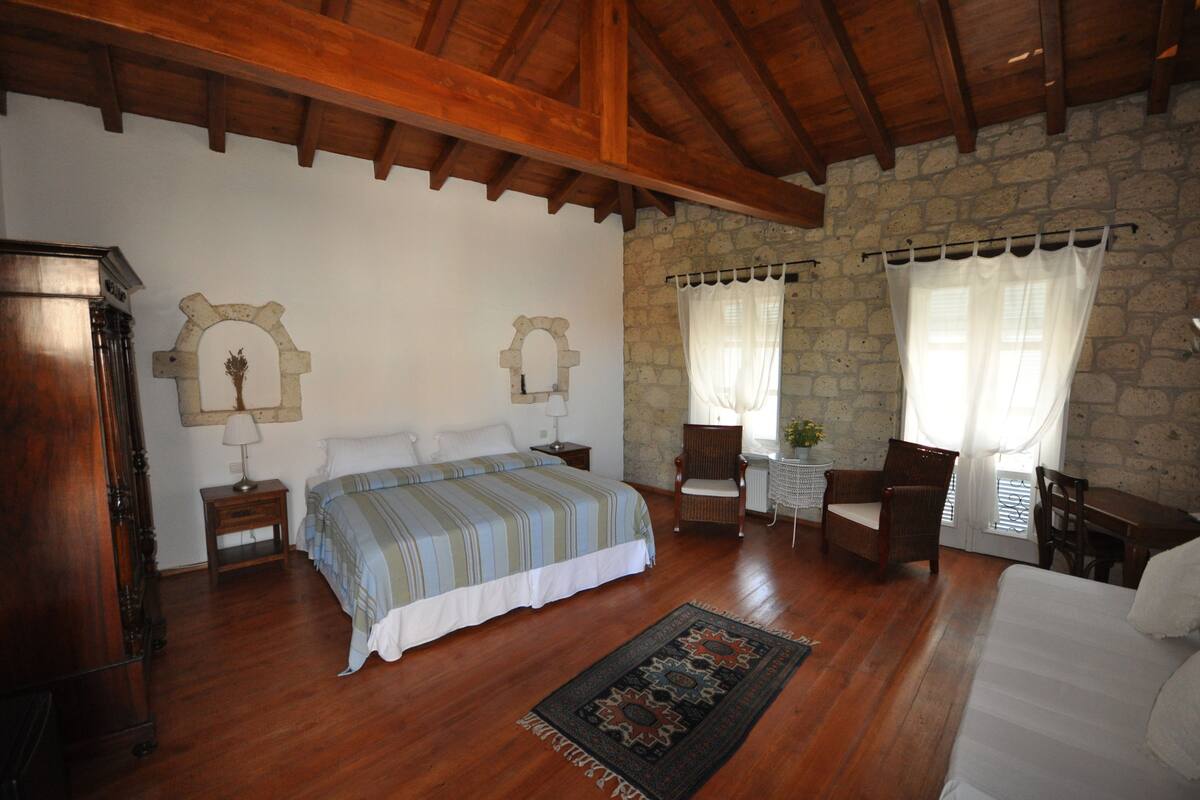 A bedroom with a bed, wooden floor, stone walls, a small table with a chair, and a window with curtains.