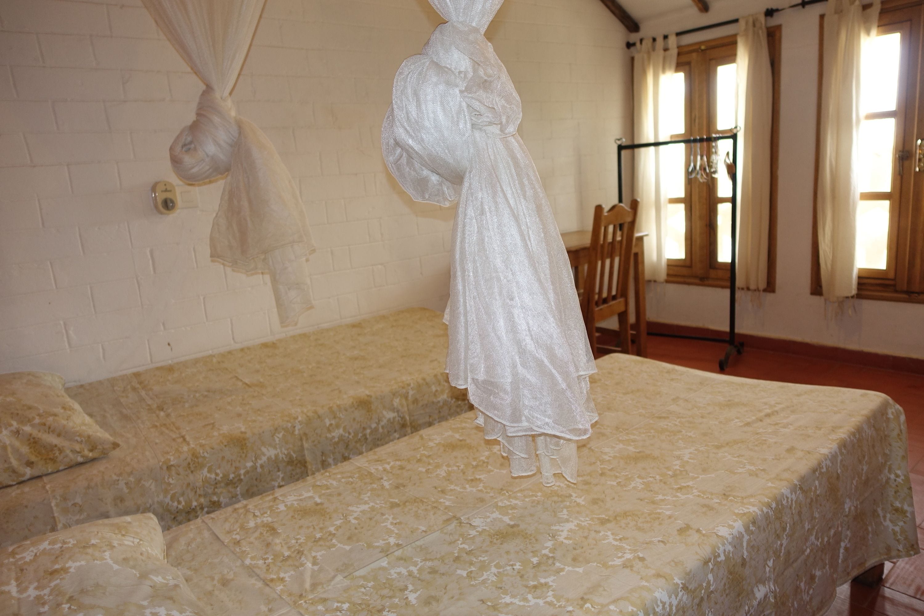 A room with two beds, a hanging white cloth, a wooden chair, and a window with curtains.