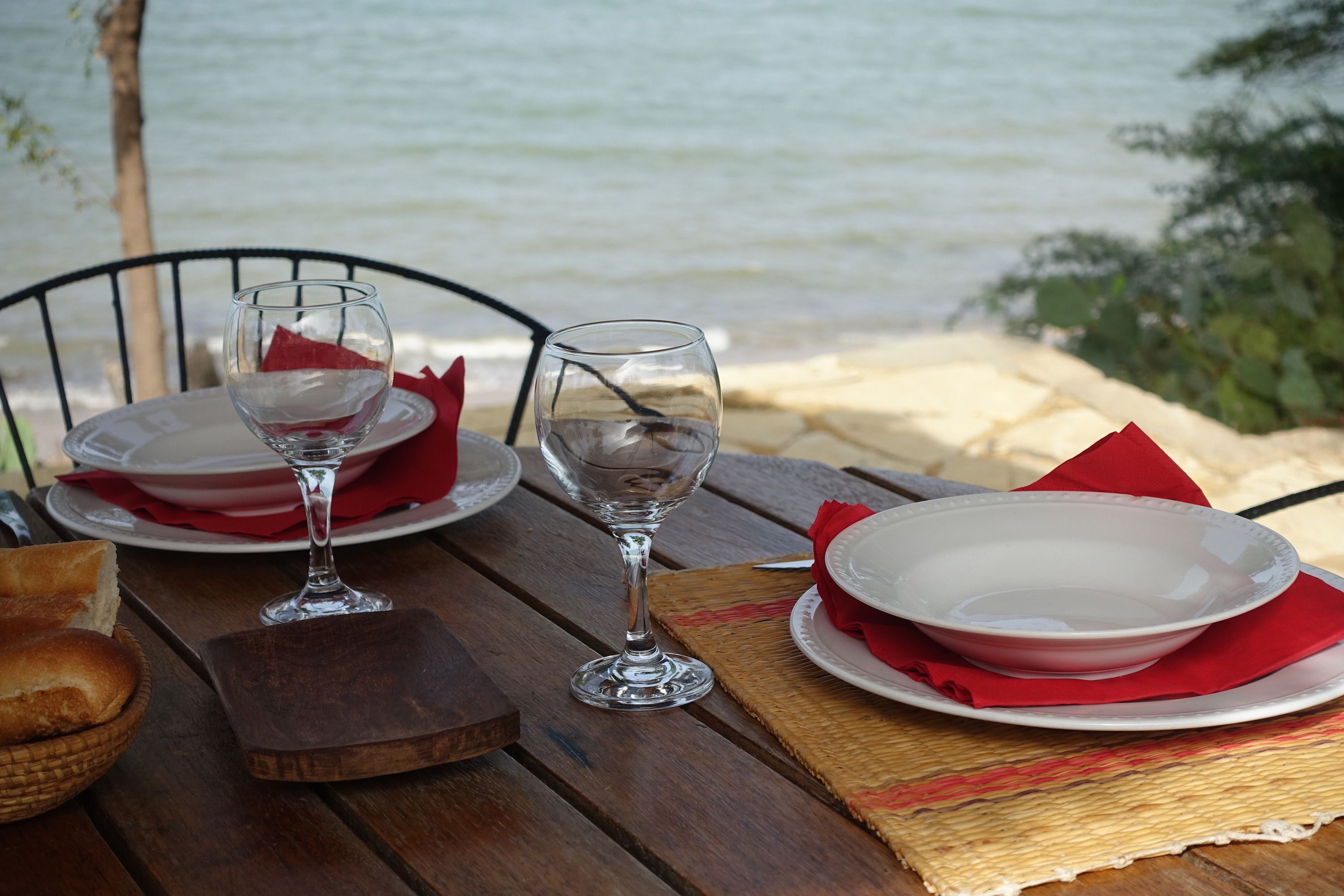 A table set with white plates, red napkins, and wine glasses, overlooking a beach.