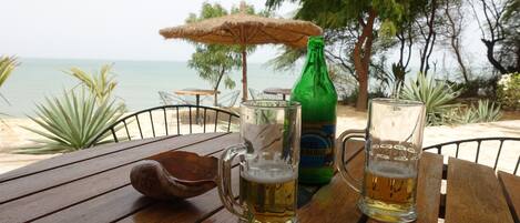 A wooden table with two beer mugs and a bottle, set outdoors with a view of the sea and trees.
