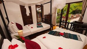 A room with two beds, each with a pillow and a blanket. There is a window with a view of greenery outside.