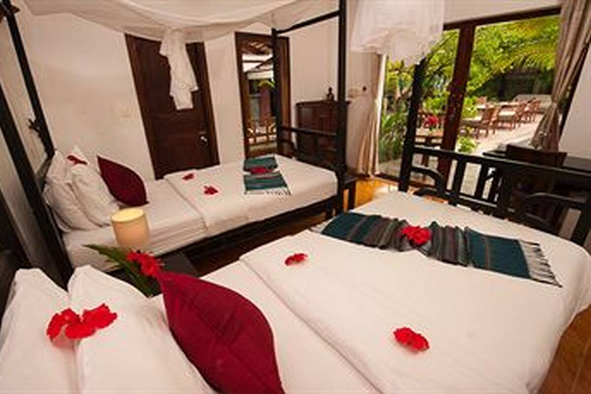A room with two beds, each with a pillow and a blanket. There is a window with a view of greenery outside.