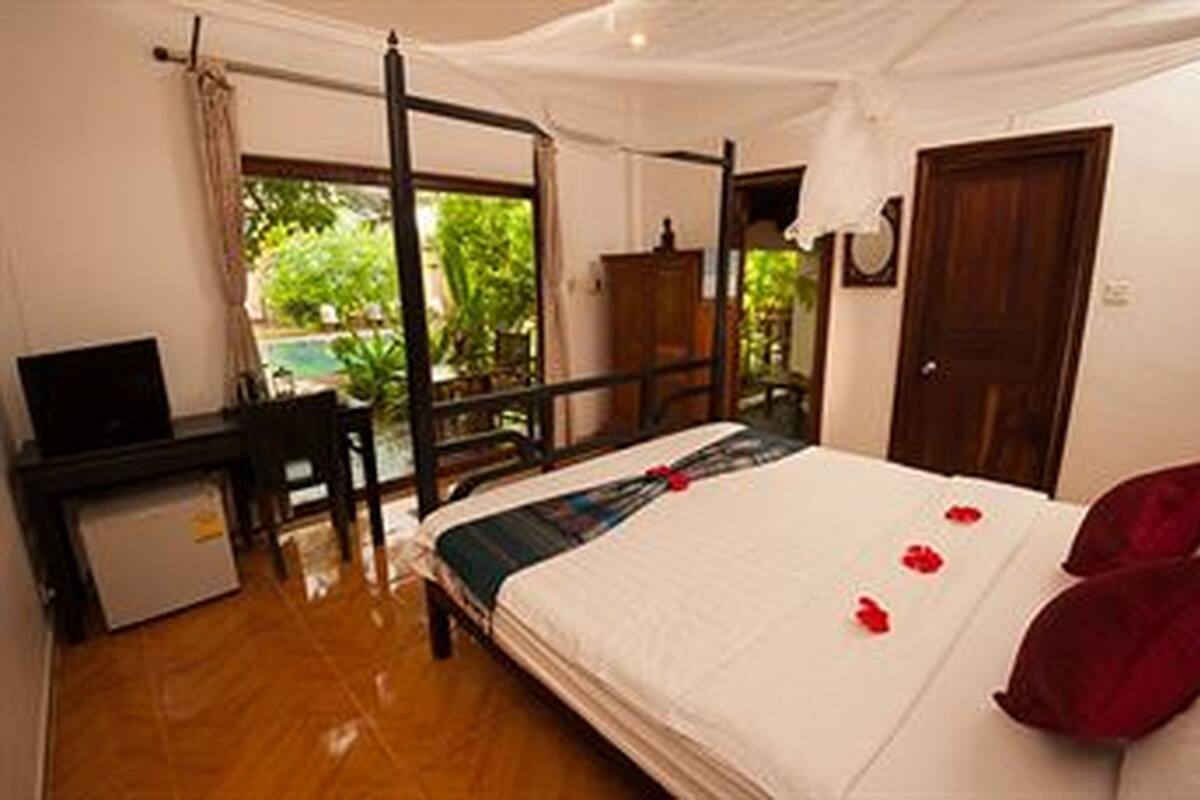 A bedroom with a four-poster bed, a small desk with a television, and a view of greenery outside.