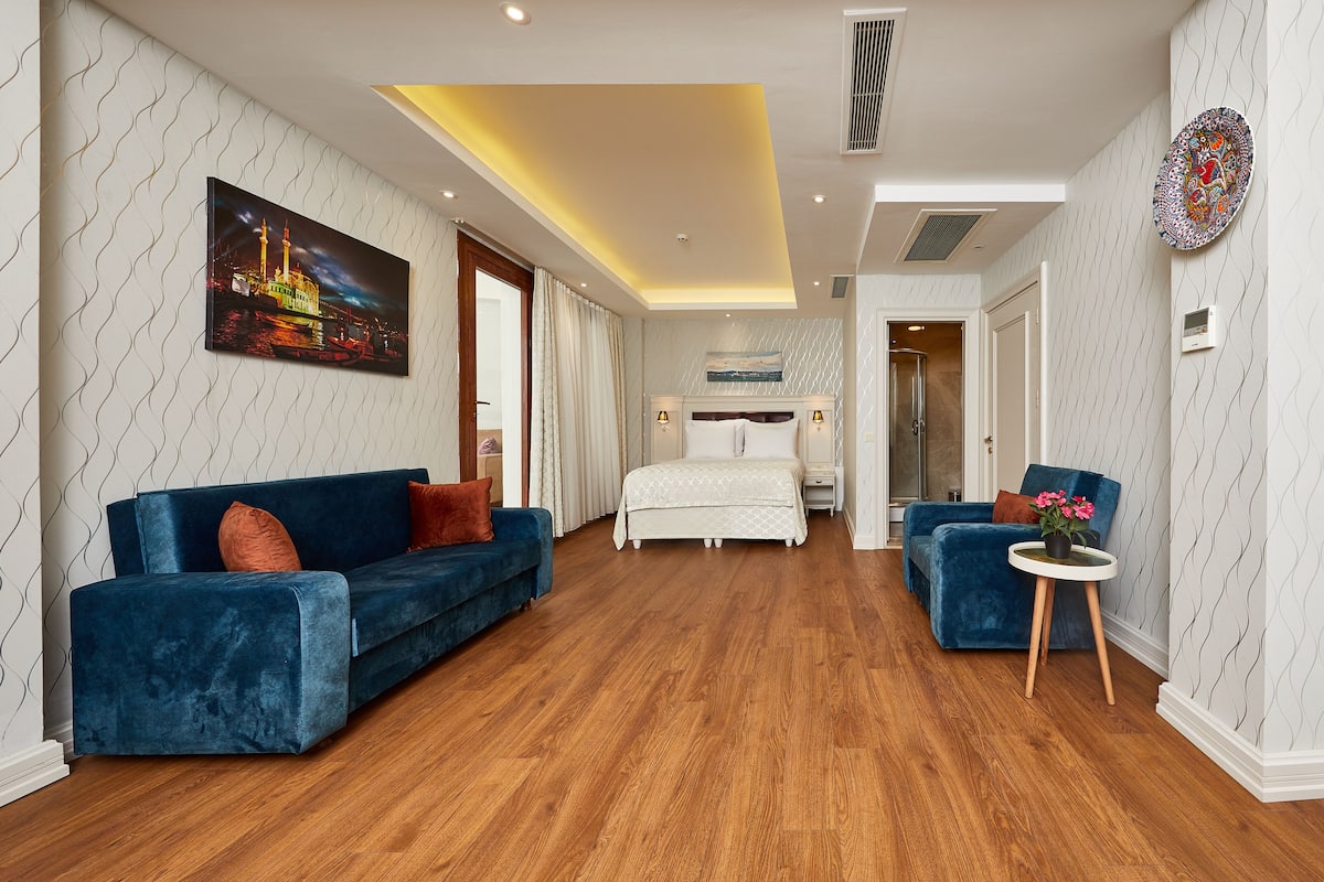Una habitación de hotel moderna con una cama grande, dos sofás azules, una mesita redonda con flores y un cuadro de un paisaje urbano en la pared.