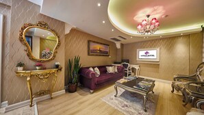 A well-lit living room with a plush purple sofa, a gold console table, and a chandelier. A television displaying the hotel name is mounted on the wall. - Albinas Hotel Old City (Istanbul)