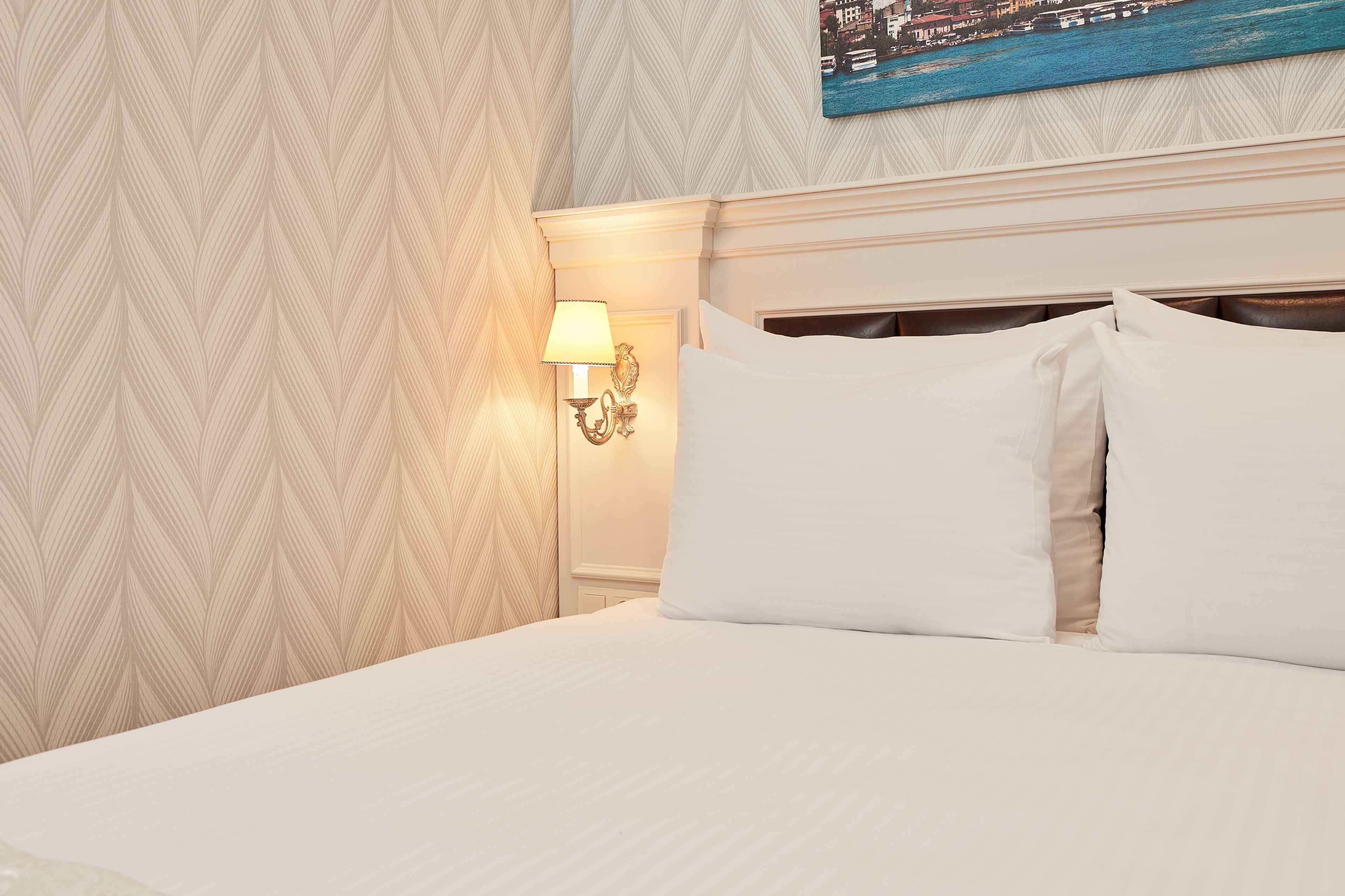 A neatly made bed with white linens in a hotel room with patterned wallpaper and a wall-mounted lamp.