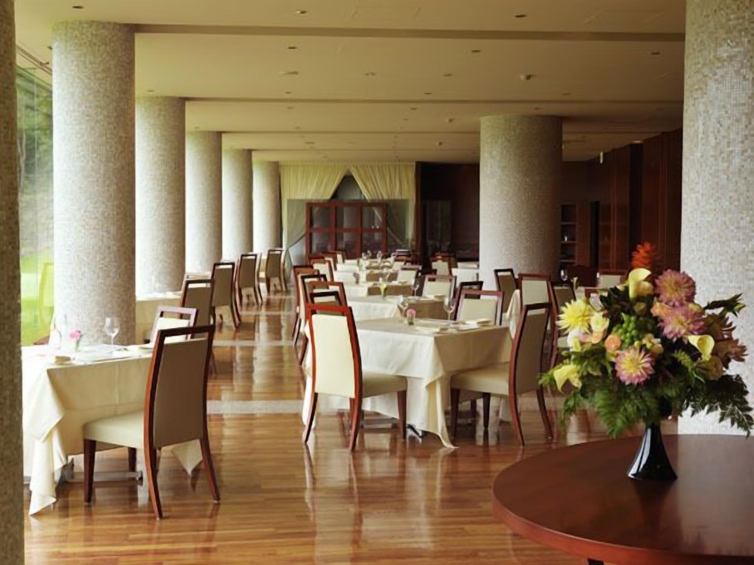 A formal dining room with tables set for multiple guests, large columns, and a view of a building in the background.