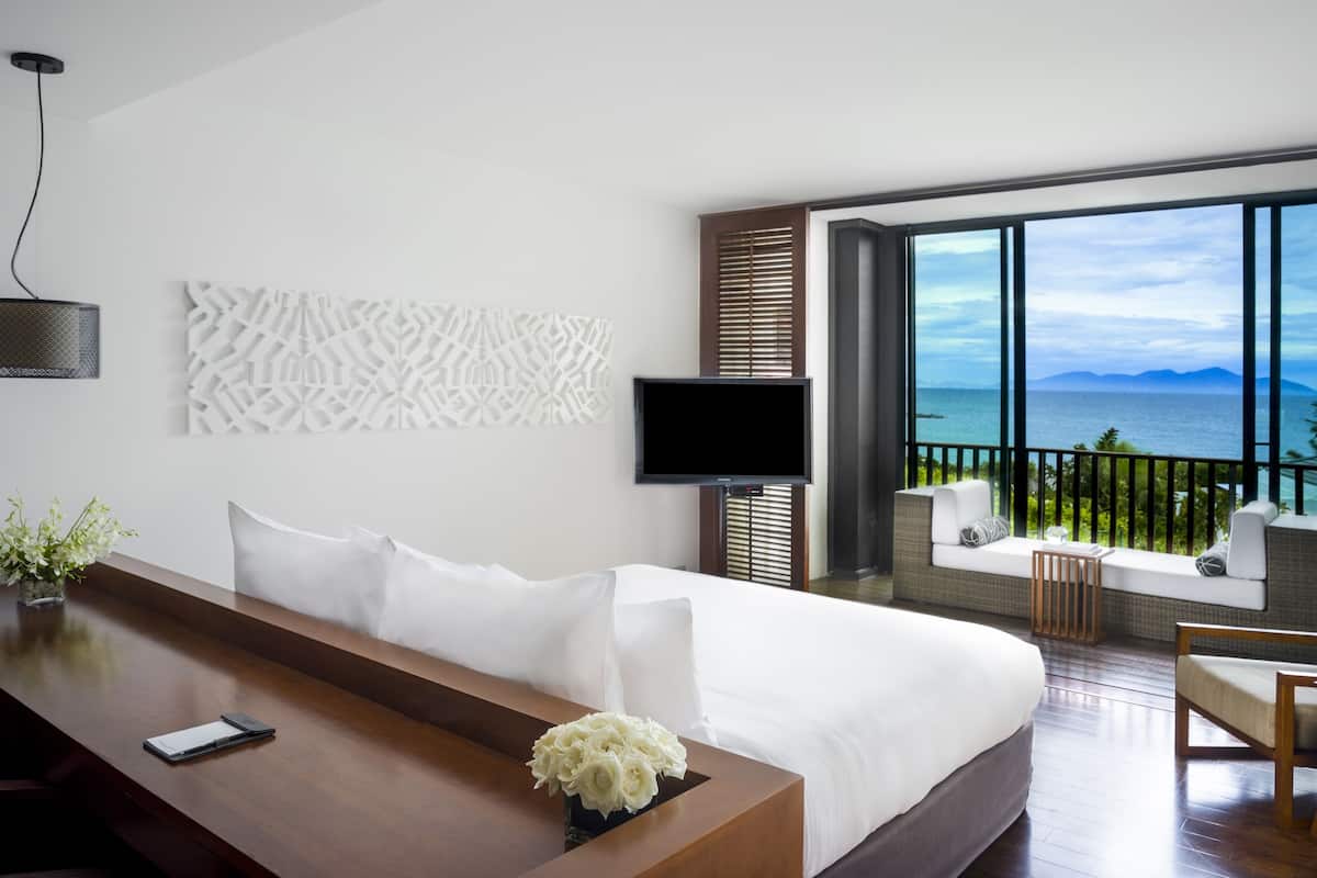 Ein modernes Hotelzimmer mit einem großen Bett, einem Fernseher, einem Schreibtisch mit Blumen und einem Balkon mit Meerblick.