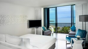 Una habitación de hotel moderna con una cama grande, televisión, balcón con vista al mar y una zona de estar con una silla y una mesa.