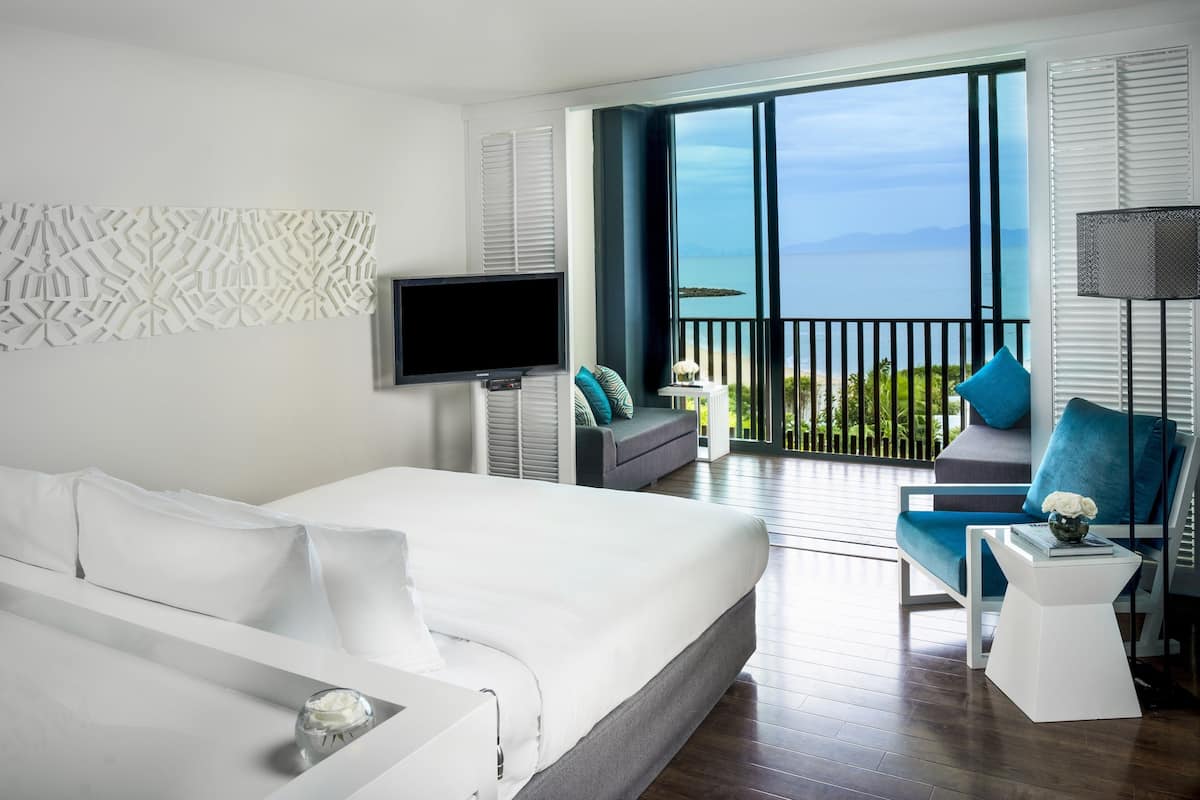 Ein modernes Hotelzimmer mit einem großen Bett, einem Fernseher, einem Balkon mit Meerblick und einer Sitzecke mit Sessel und Tisch.
