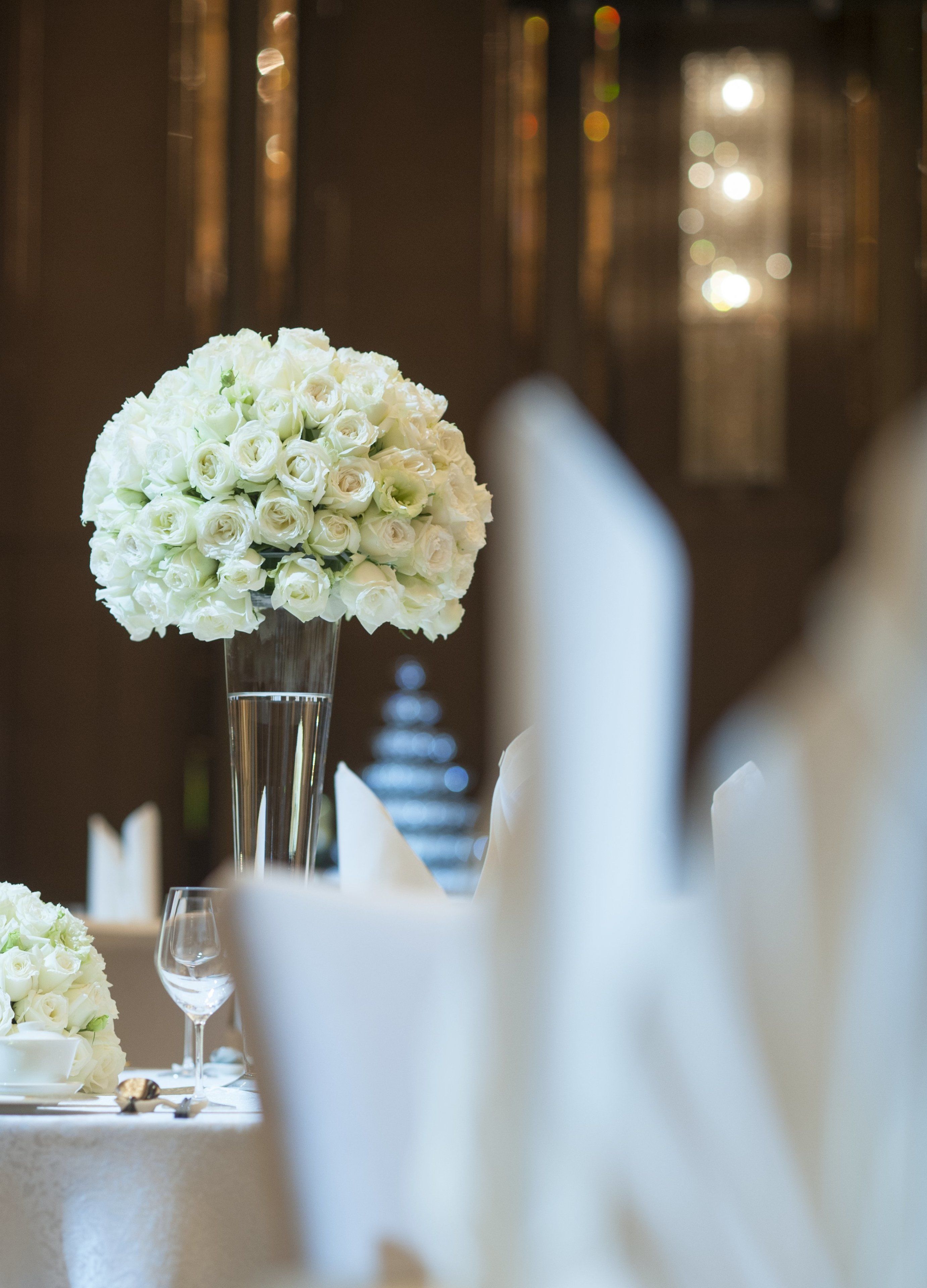 Una mesa con un florero alto de vidrio con flores blancas, un vaso de agua y una servilleta doblada.