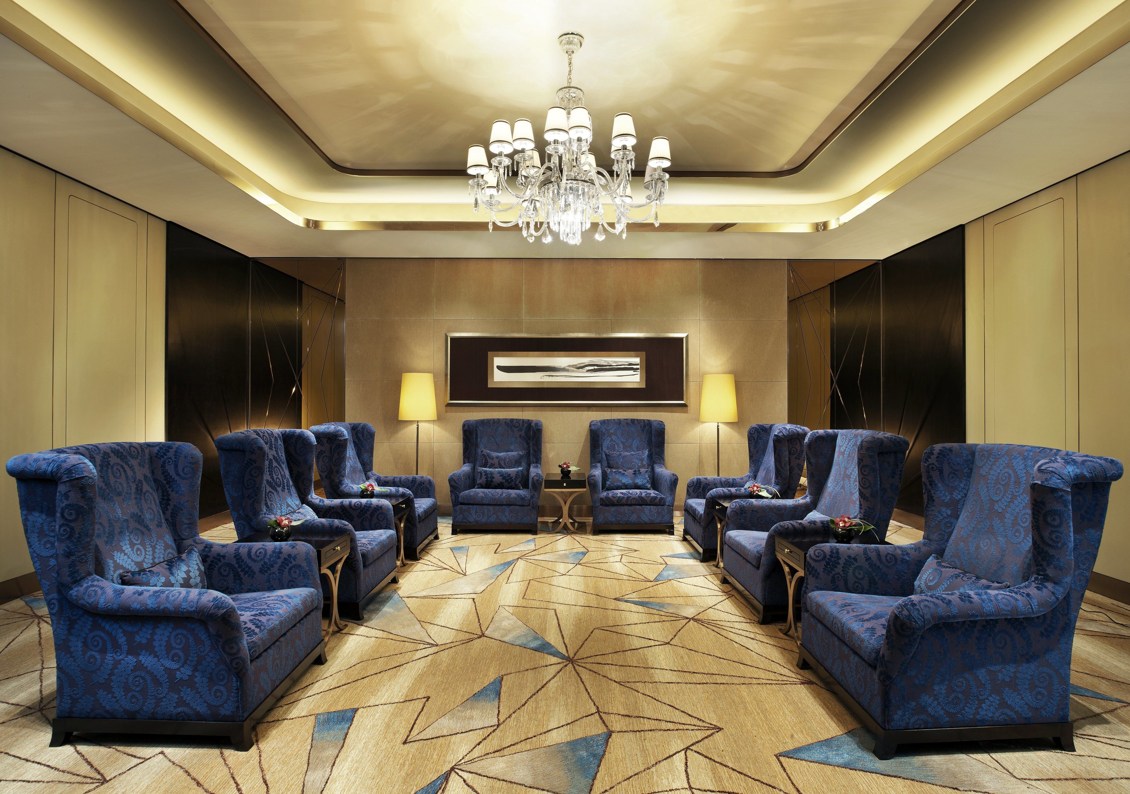 A hotel lobby with a geometric patterned carpet, a chandelier, and blue patterned armchairs arranged around a central table.