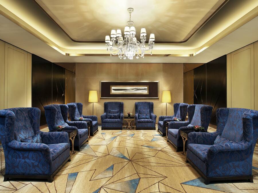 Un vestíbulo de hotel con alfombra de estampado geométrico, un candelabro y sillones azulados de diseño dispuestos alrededor de una mesa central.