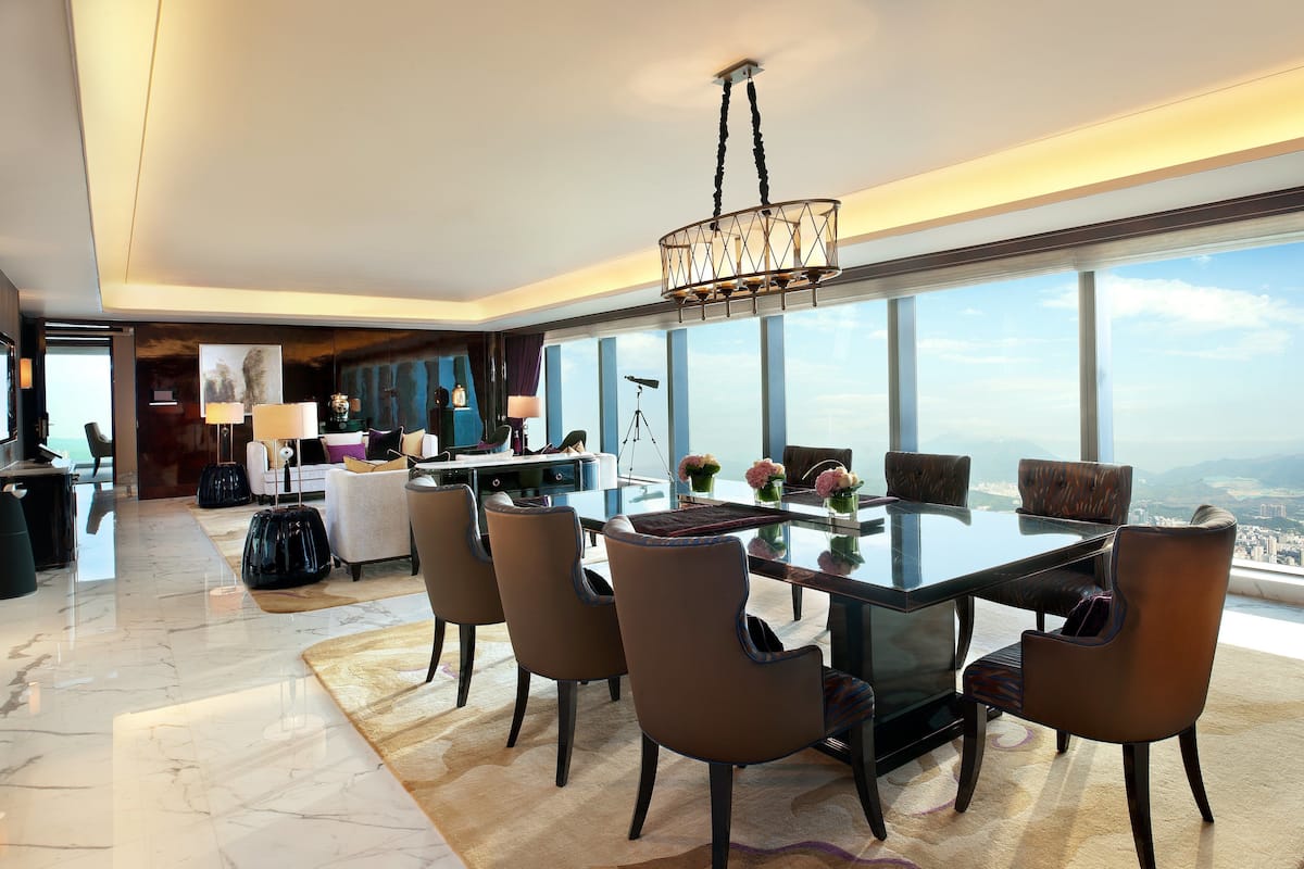 A modern hotel room with a large dining table, comfortable seating, a chandelier, and a city view.