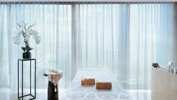 A modern spa room with a large window, a massage table, a small side table with a vase of flowers, and a sink area.
