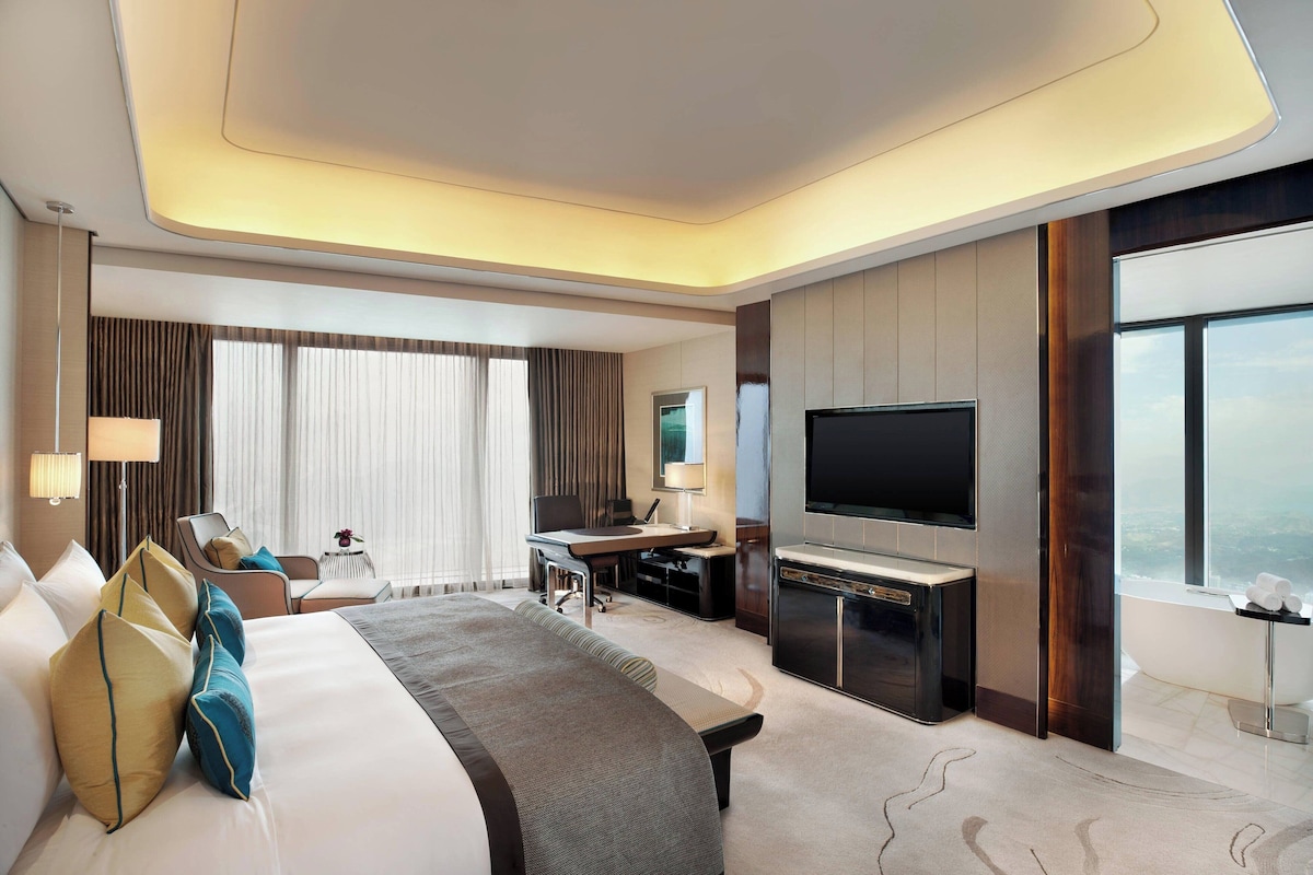A hotel room with a large bed, a desk, a flat-screen TV, and a view of the city.