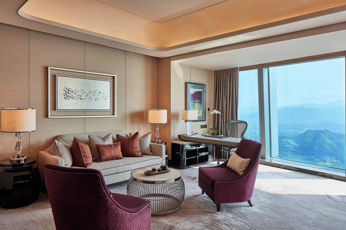 Una habitación de hotel moderna con sofá, dos sillones, una mesa de centro y un escritorio con lámpara. Un ventanal grande ofrece vistas a la ciudad.