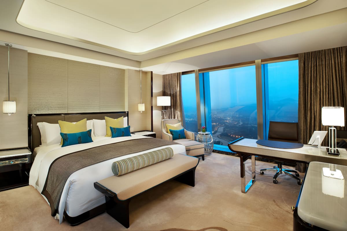 A hotel room with a large bed, a desk, a chair, a bench, a view of the city, and a large window.