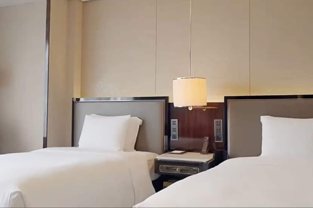 A hotel room with two beds, a nightstand with a lamp, and a control panel.