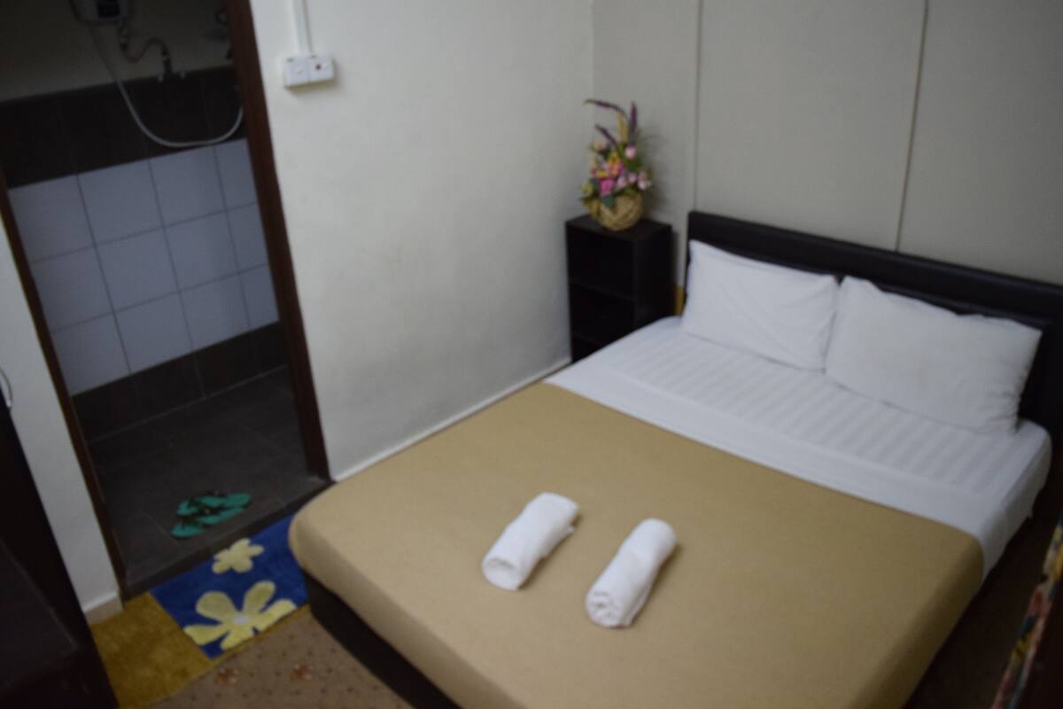 A hotel room with a bed, a small bedside table with a vase of flowers, a shower area, and a bathroom with a towel rack.