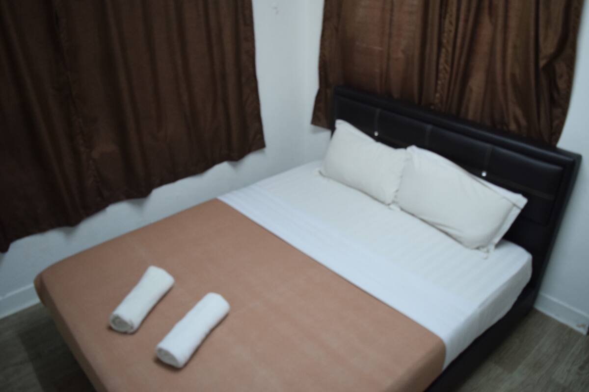 A single bed with a brown headboard, white bedding, and two rolled towels on a brown bedside table.