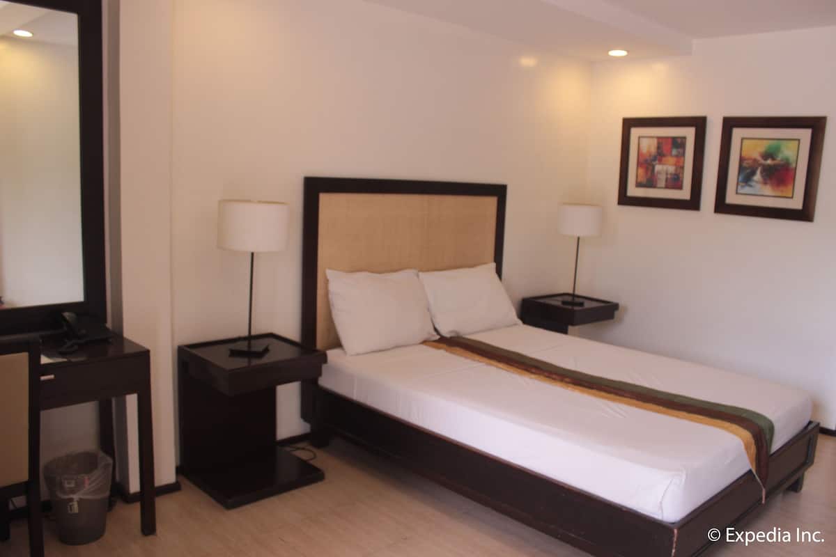 Quarto de hotel com uma cama de solteiro, duas mesas de cabeceira com luminárias, uma escrivaninha com cadeira e um cesto de lixo.