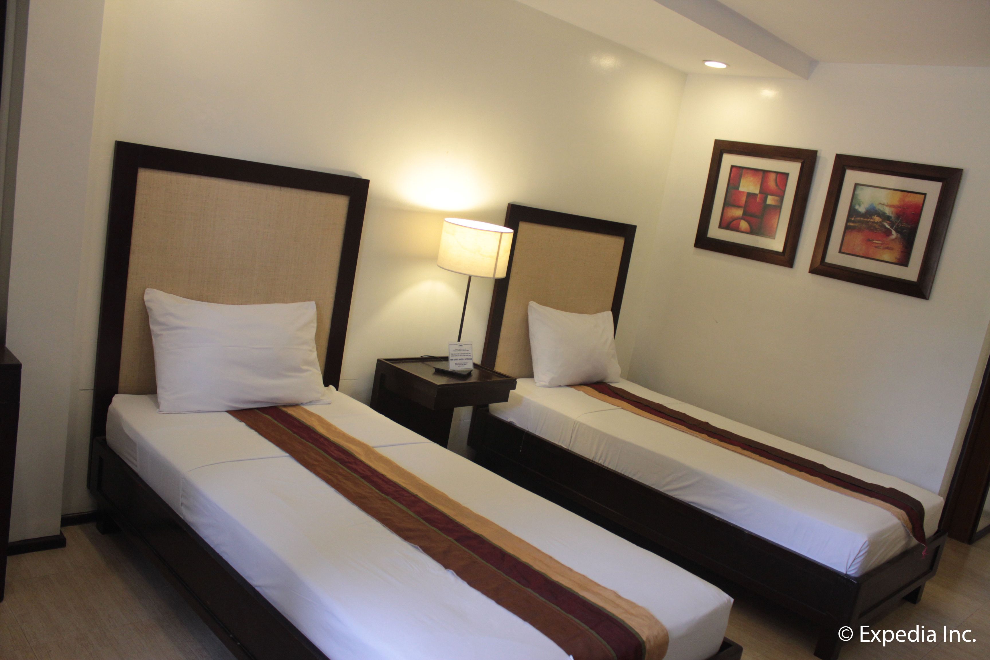 A hotel room with two single beds, each with a wooden headboard and white bedding. There is a nightstand with a lamp between the beds, and two framed pictures on the wall.