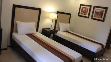 A hotel room with two single beds, each with a wooden headboard and white bedding. There is a nightstand with a lamp between the beds, and two framed pictures on the wall.