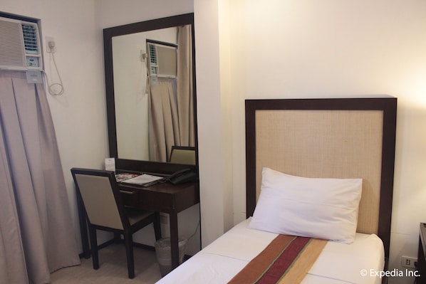 A hotel room with a bed, a desk, and a chair. There is a mirror on the wall, and an air conditioning unit is mounted on the wall.