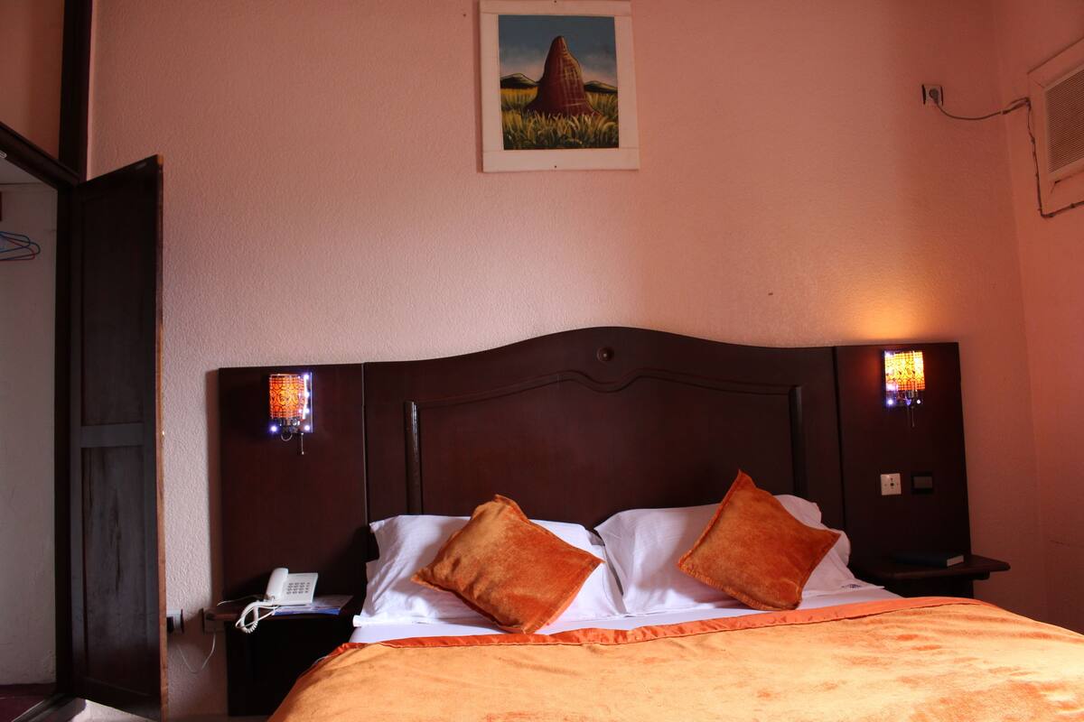 A hotel room with a wooden headboard, two pillows, a bedside table with a phone, and a painting on the wall.