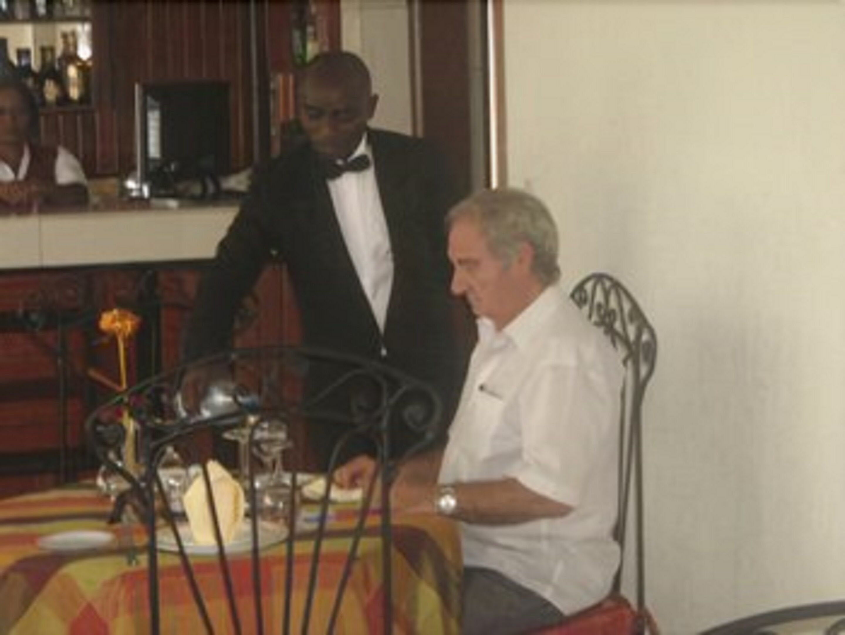 A dining area with a table set for two, a bar in the background, and a waiter attending to a guest.