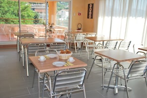 A dining area with multiple tables and chairs, a basket of croissants, and a view of greenery through large windows.