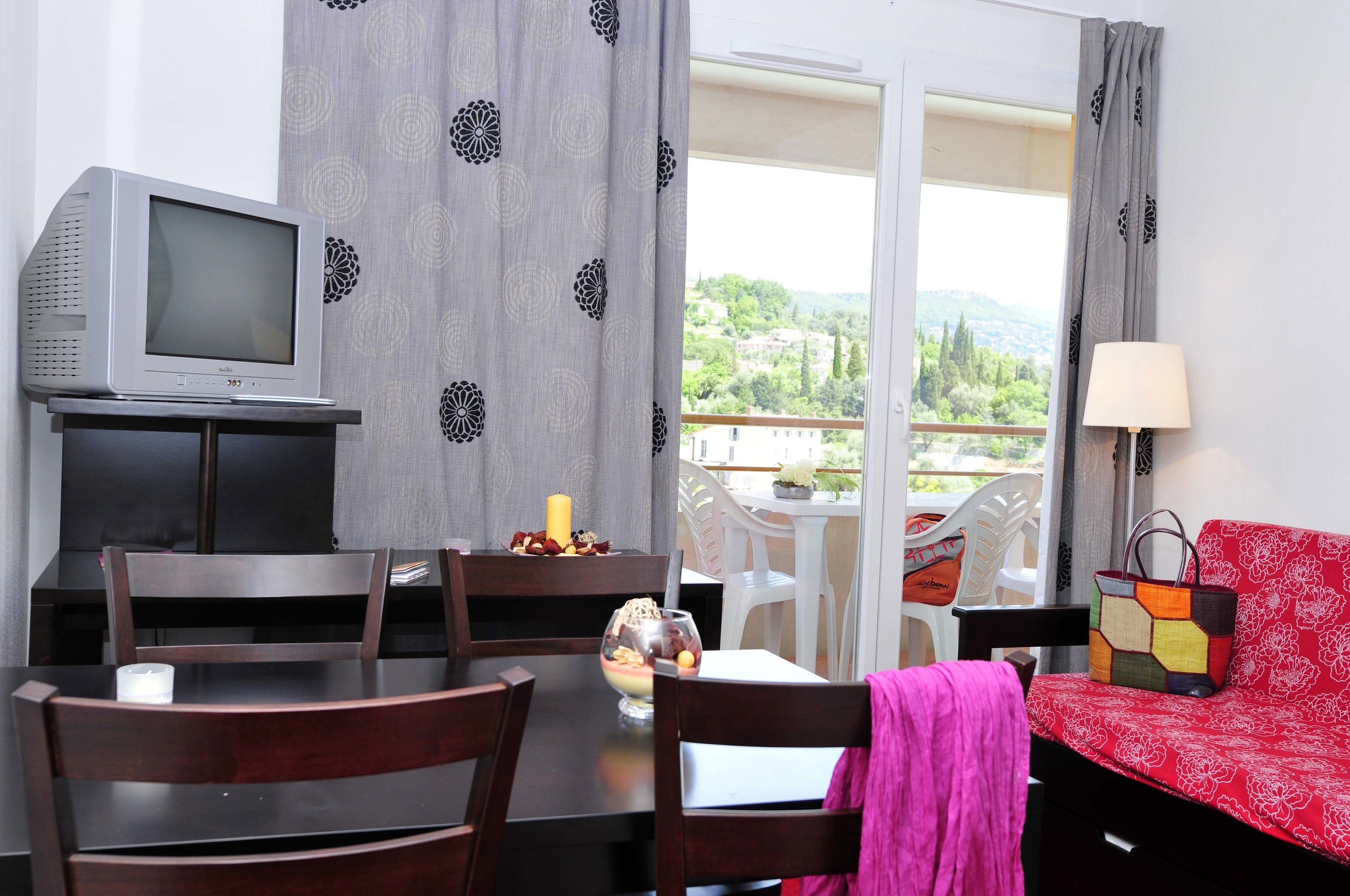 A room with a dining table, chairs, a TV, a sofa, a balcony with a view, and a lamp.