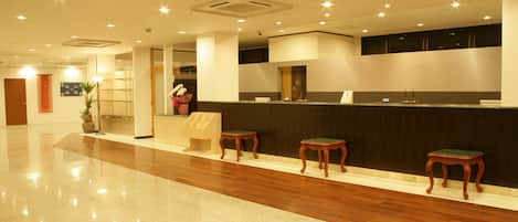 Zona de recepción de hotel con suelo pulido, mostrador de recepción y tres taburetes de madera.
