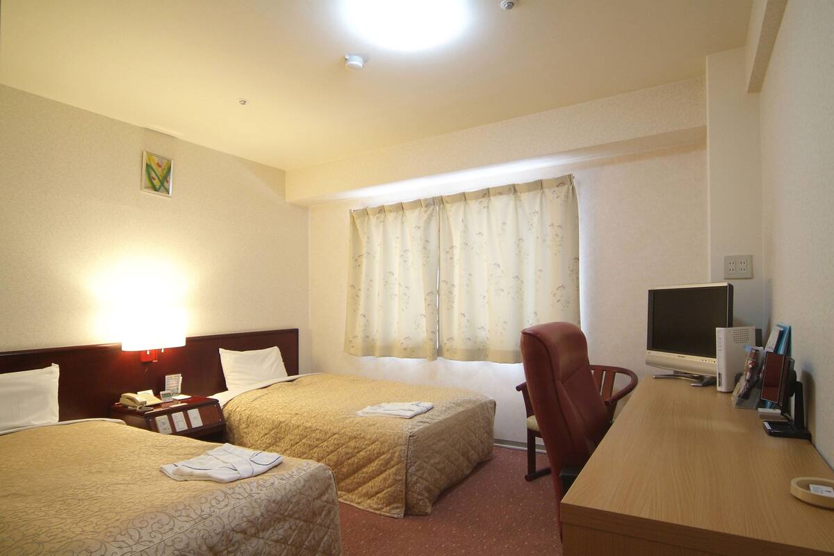 Una habitación de hotel con dos camas, un escritorio con computadora, una silla y un ventanal con cortinas.