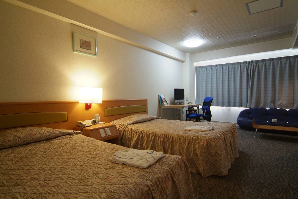 Habitación de hotel con dos camas, un escritorio con computadora, una silla y un ventanal grande con cortinas.