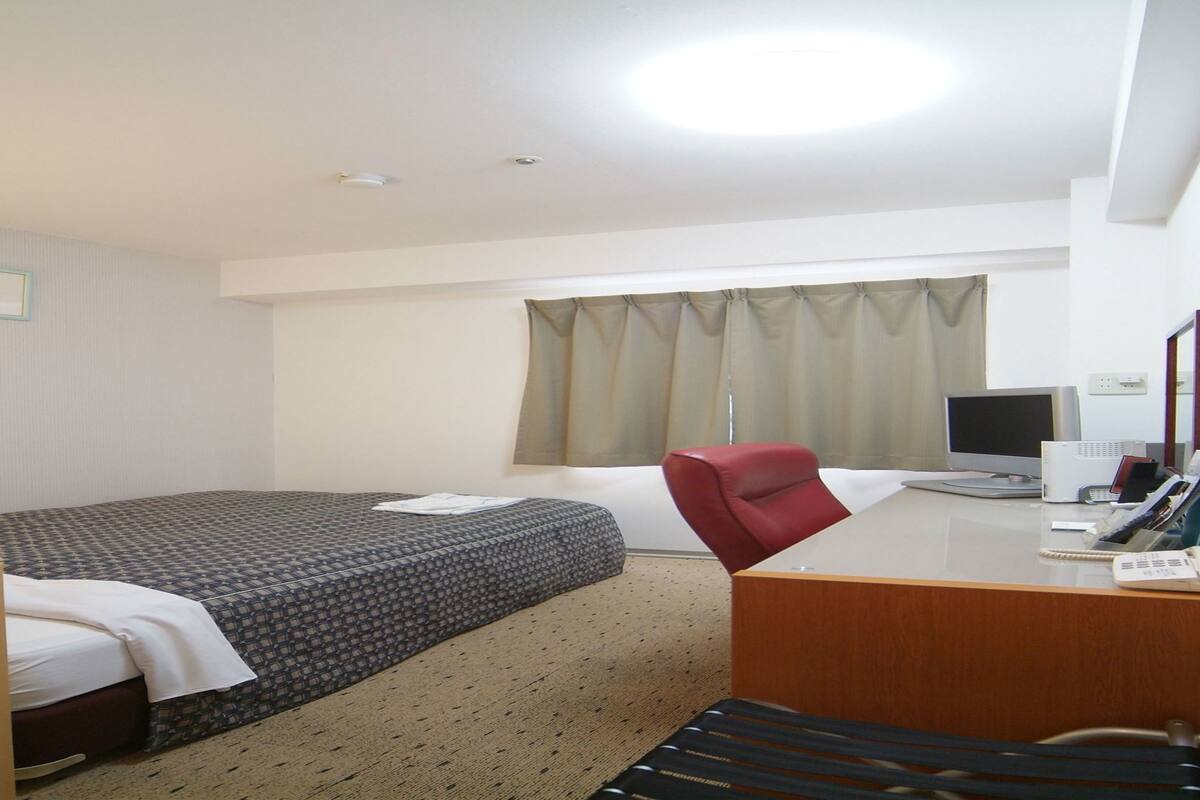 Habitación de hotel con cama, escritorio con computadora, una silla y ventana con cortinas.