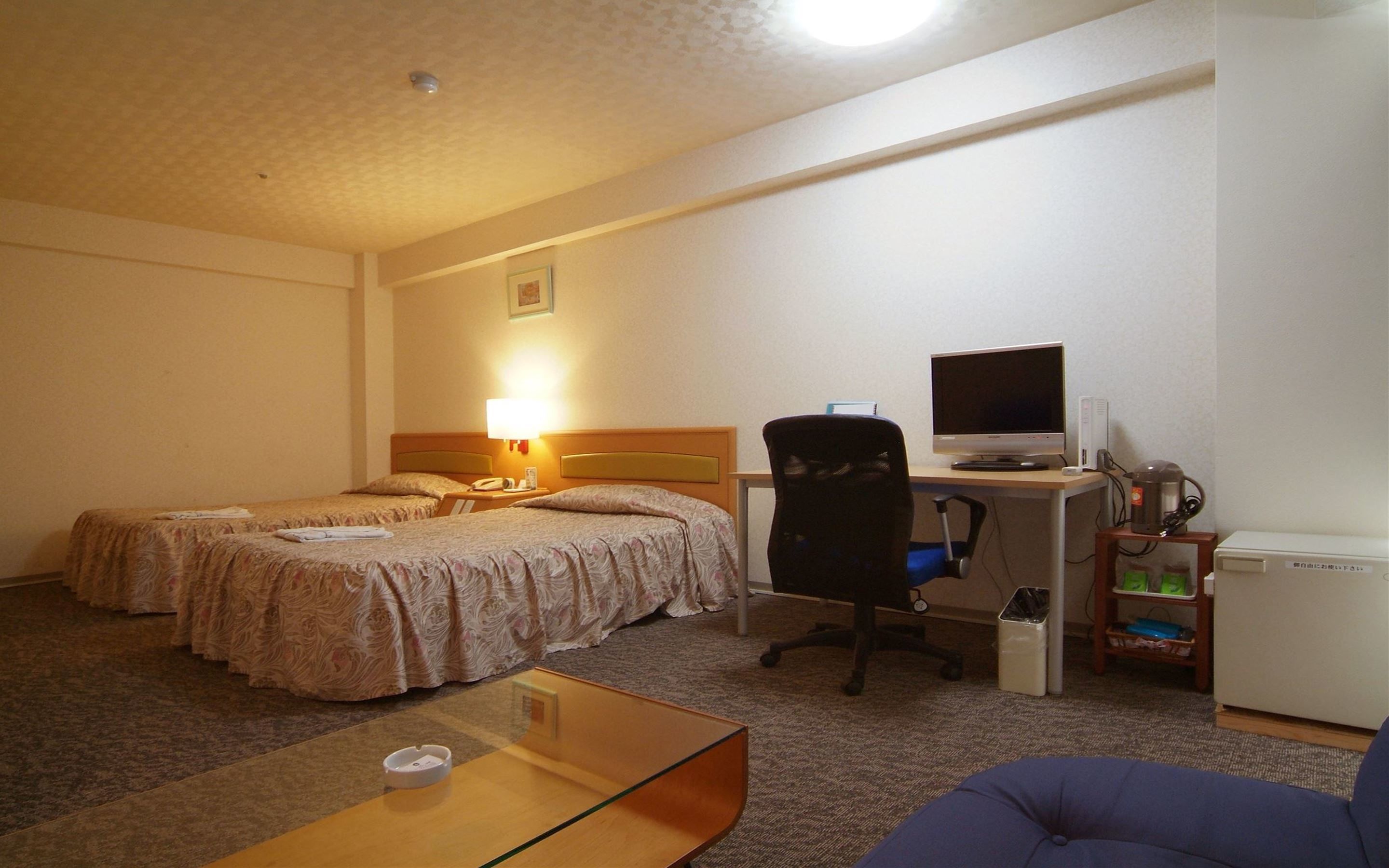 A hotel room with two beds, a desk with a computer, a chair, a small table, and a trash bin.