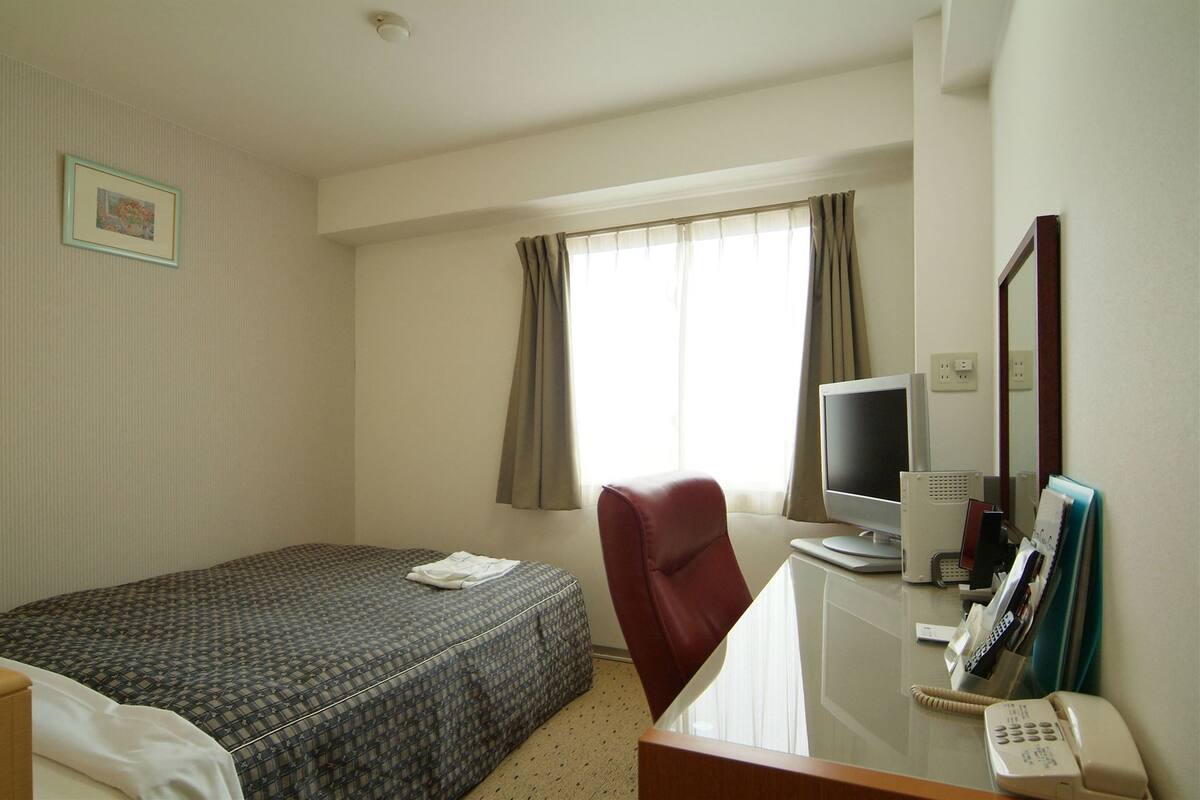 Habitación de hotel con cama, escritorio, computadora, teléfono y ventana con cortinas.