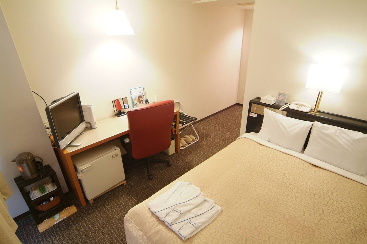 Una habitación de hotel con una cama, un escritorio con computadora, una silla, un refrigerador pequeño y una mesita de noche con lámpara.
