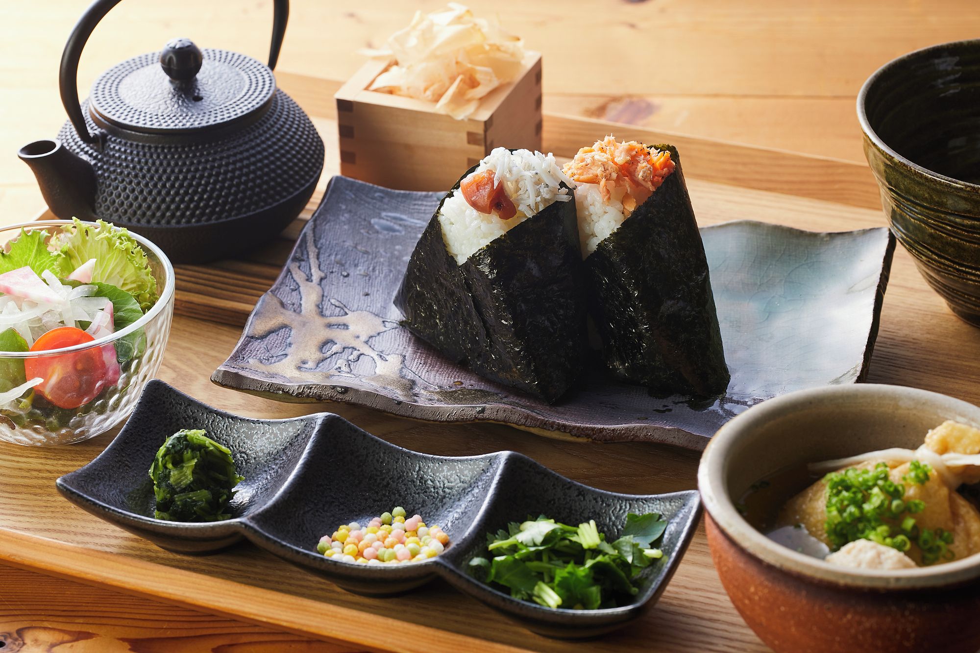 Een houten tafel met een zwarte theepot, een saladekom en diverse gerechten, waaronder sushi.