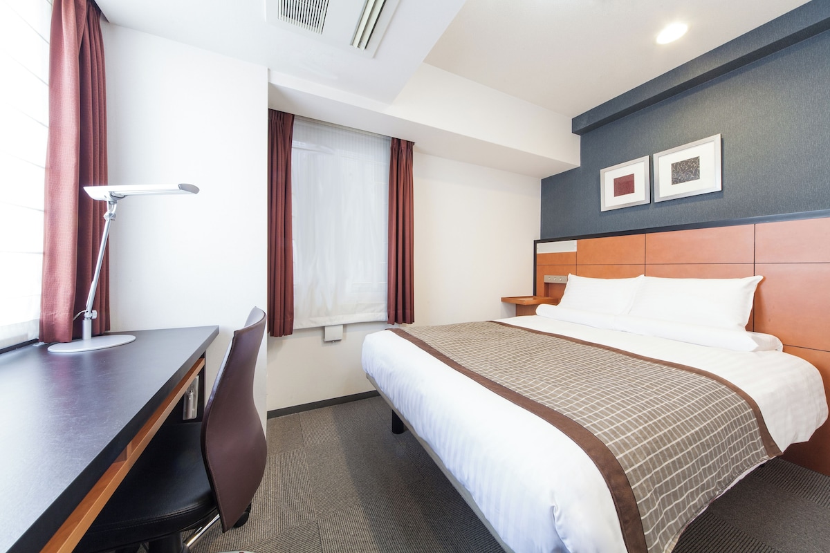 Een hotelkamer met een bed, een bureau, een stoel, een raam met gordijnen en twee ingelijste foto's aan de muur.
