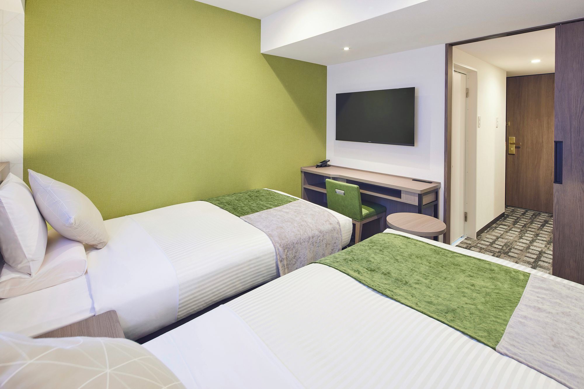 A hotel room with two beds, a desk, a chair, a flat-screen TV, and a door leading to another room.