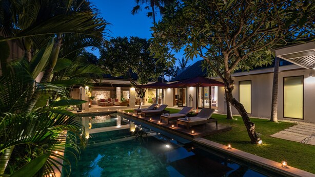 A swimming pool with lounge chairs, a dining area, and a well-lit building. - Chandra Bali Villas (Seminyak)